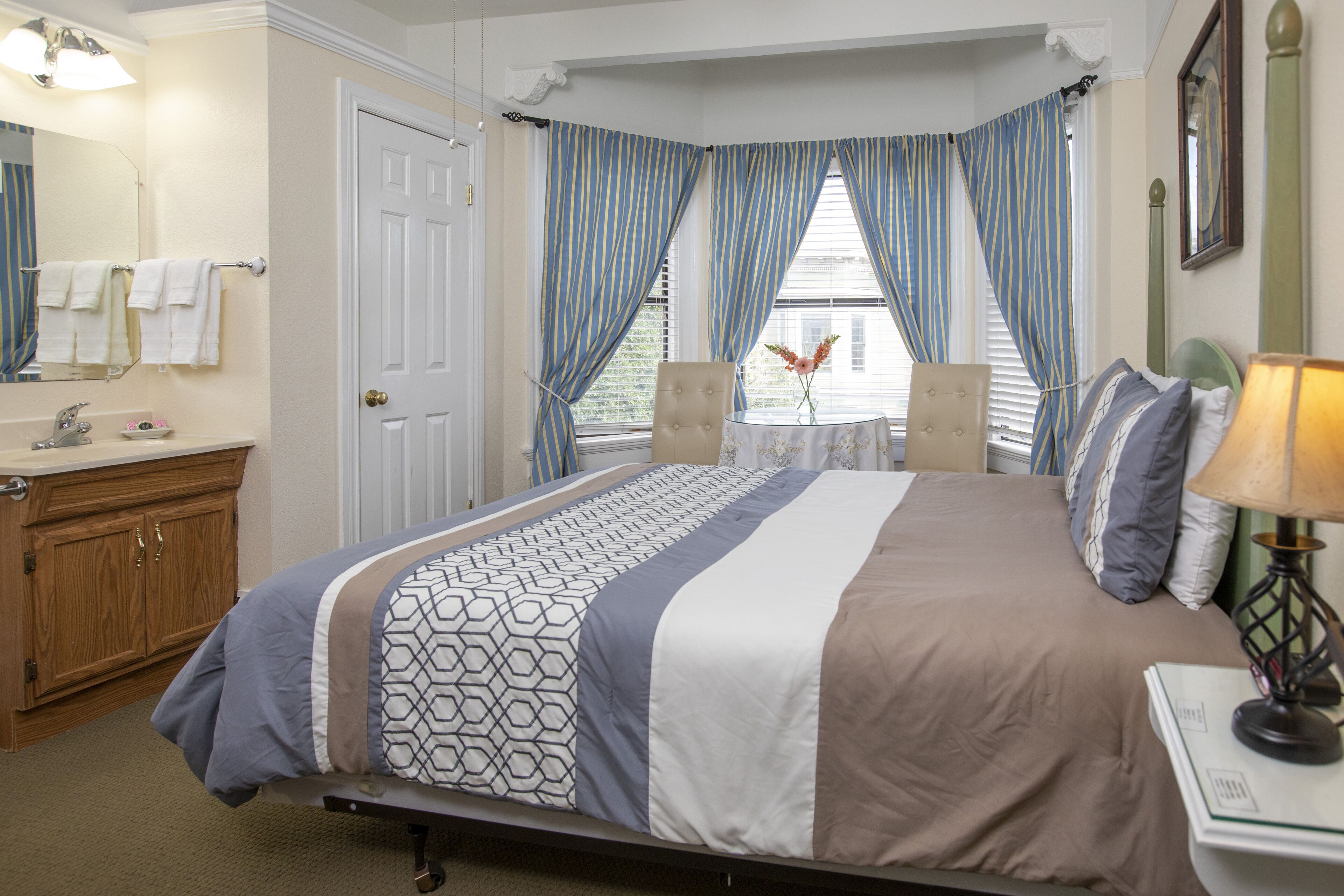 Quarto com cama, mesa de cabeceira, luminária, cadeira, janela com cortinas, espelho e banheiro com pia e porta-toalhas.