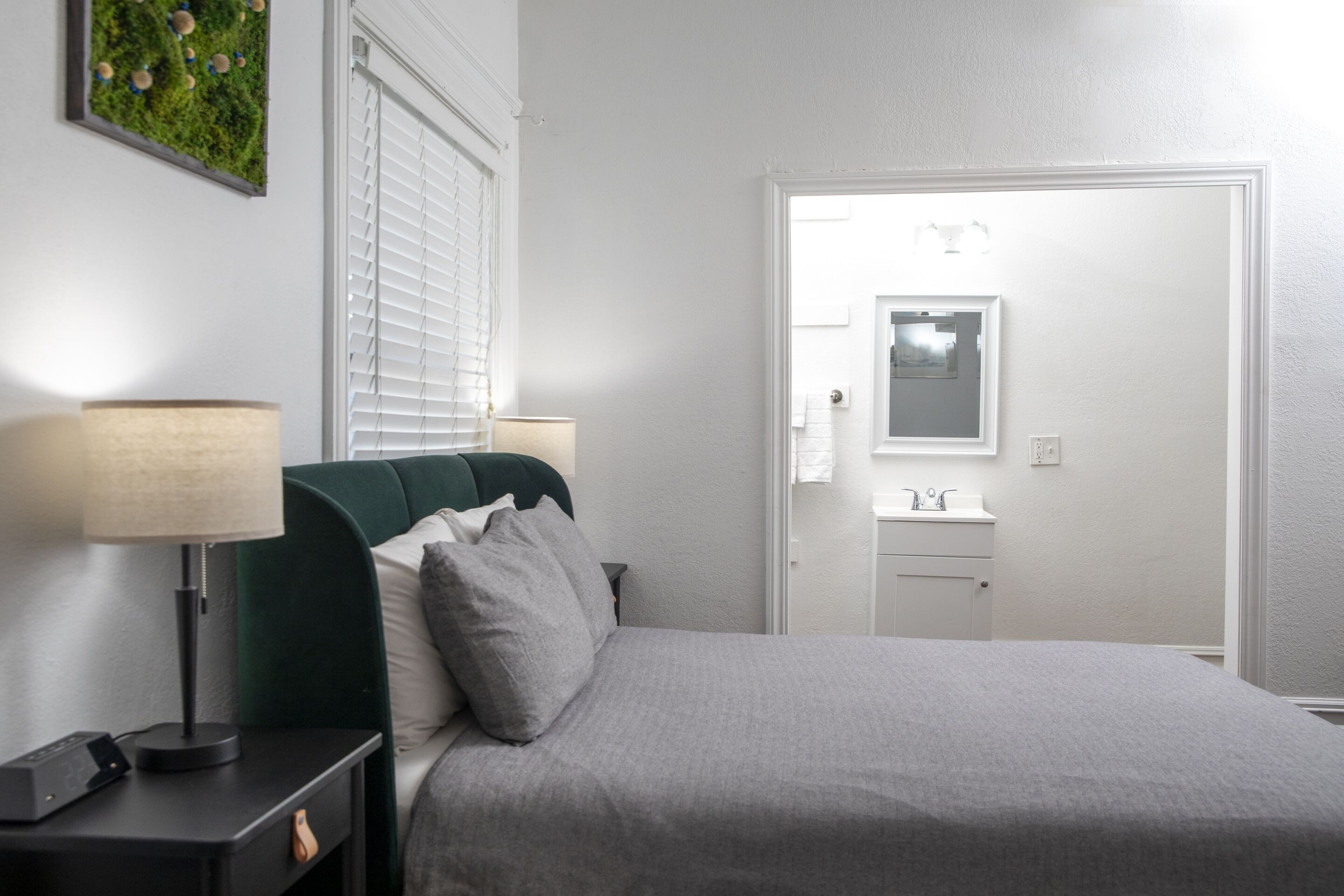 A neatly arranged bedroom with a bed, bedside lamps, a nightstand, a window with blinds, and a bathroom visible through an open door.