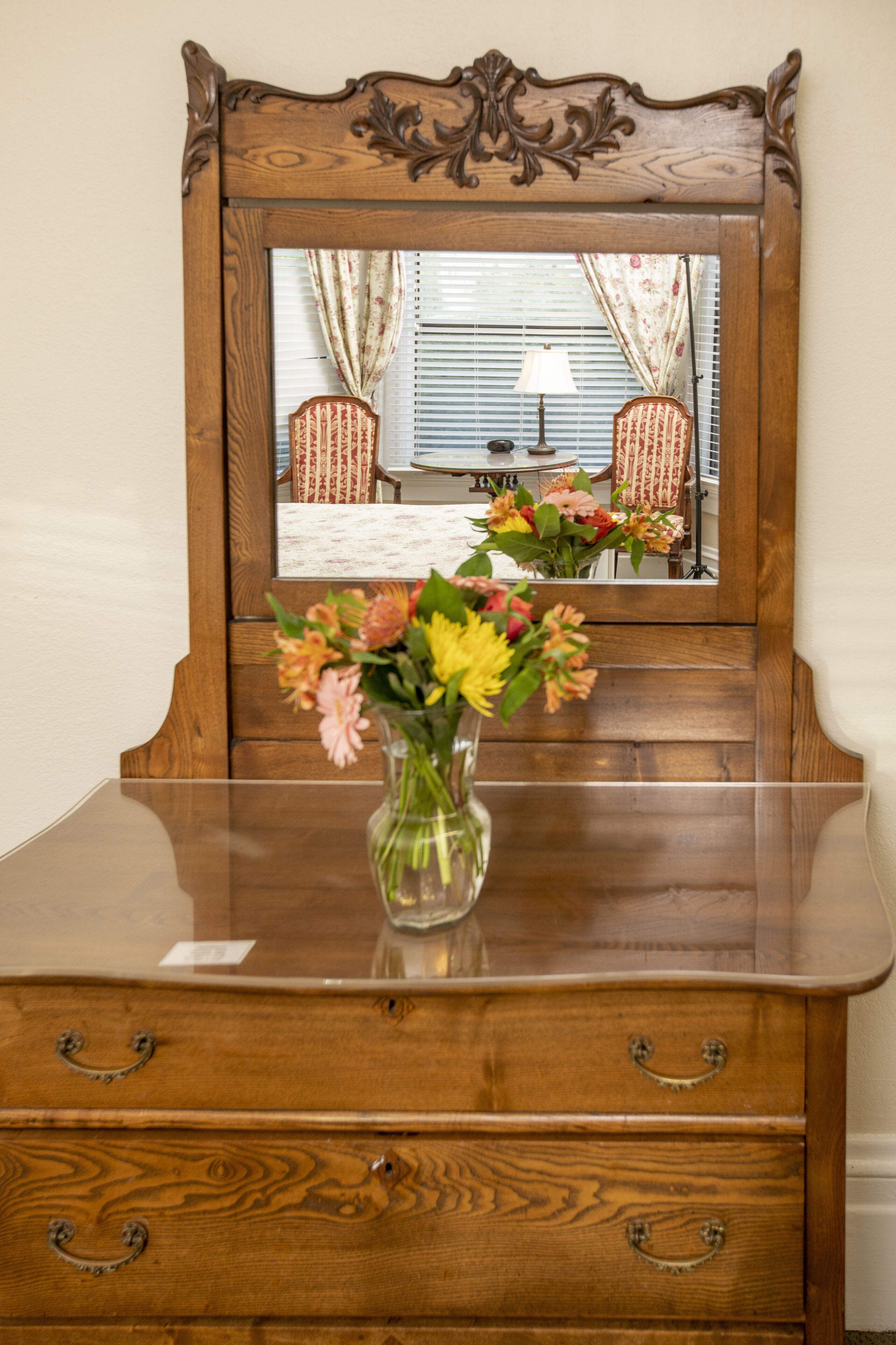 Un aparador de madera con un espejo arriba que refleja una habitación con una ventana, cortinas y una lámpara.