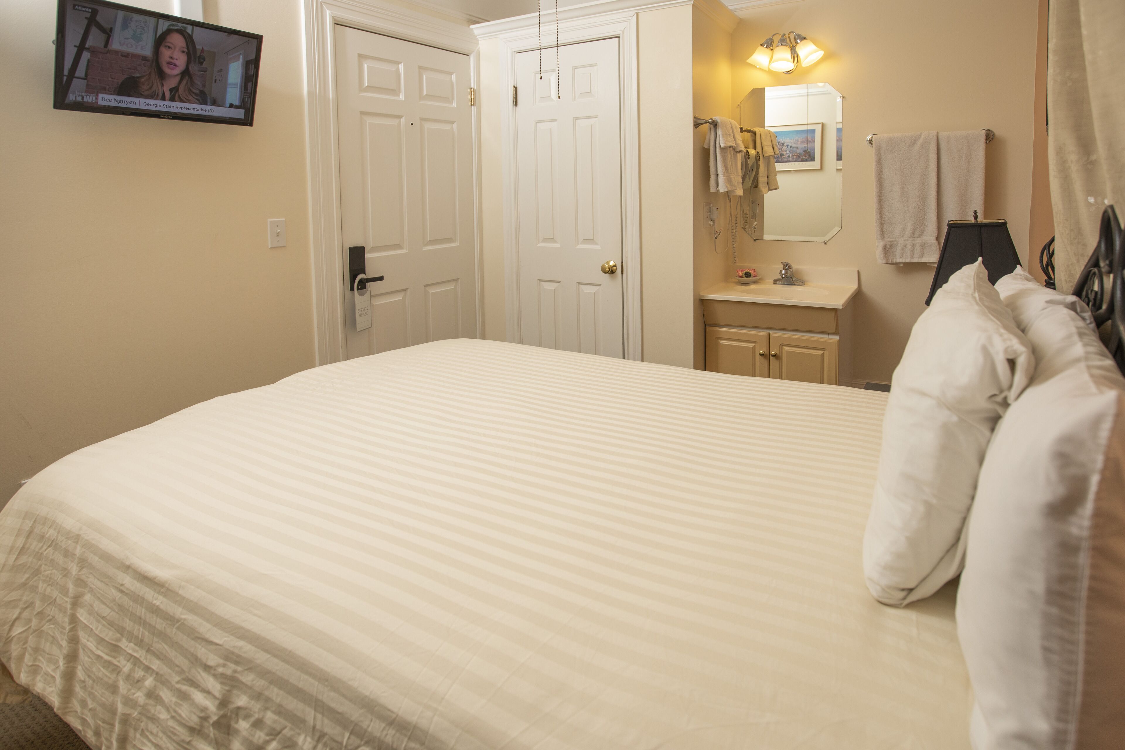 A neatly made bed with a striped bedspread, a white door, a bathroom with a sink and mirror, and a television mounted on the wall.