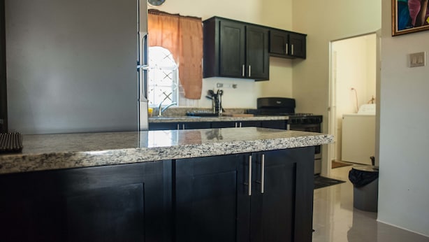 A kitchen with dark cabinets, a granite countertop, a refrigerator, and a clock on the wall. - Island Blues Cottage at Draxhall Country Club (Ocho Rios)
