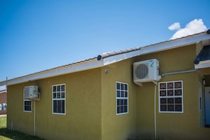 A yellow building with two air conditioning units mounted on the exterior wall, a tiled roof, and a small window with a grid pattern. - Island Blues Cottage at Draxhall Country Club (Ocho Rios)