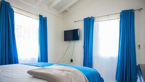 A hotel room with a bed, a TV mounted on the wall, and blue curtains. - Island Blues Cottage at Draxhall Country Club (Ocho Rios)