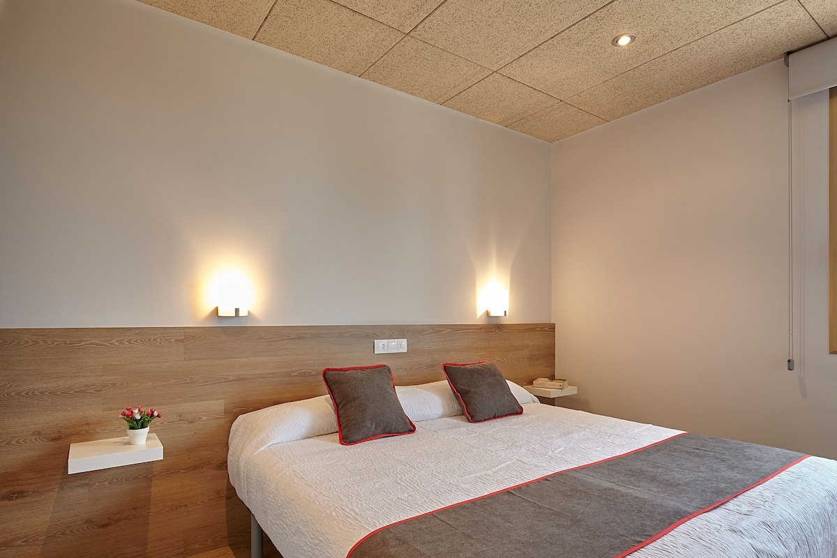 A neatly arranged bedroom with a wooden headboard, a bed with a white and grey bedspread, two bedside tables with lamps, and a small plant on one table.