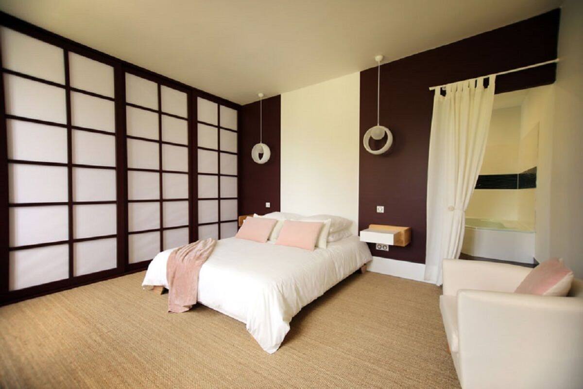 A bedroom with a bed, a chair, a bedside table, a window with curtains, and a wall with a traditional Japanese screen.