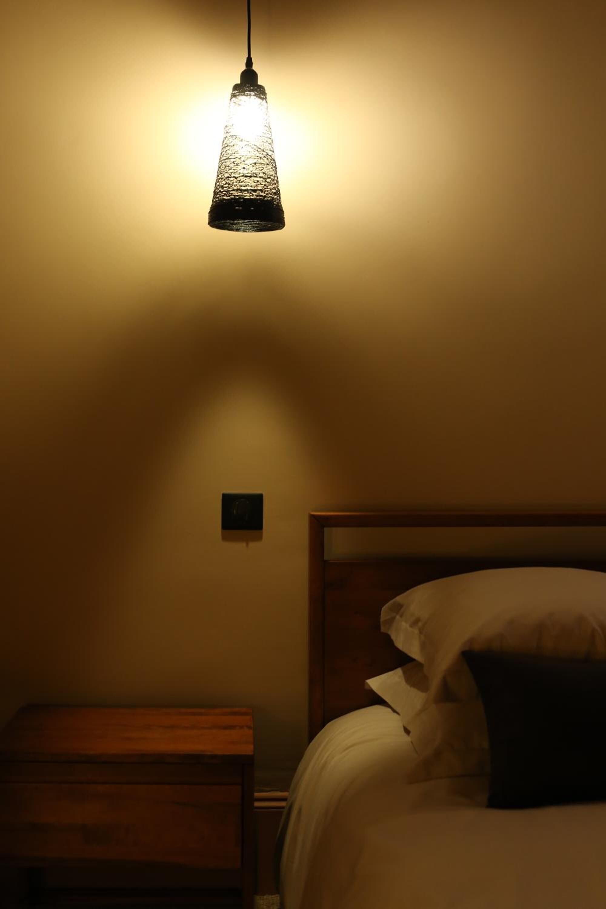 Une pièce faiblement éclairée, avec un lit, une table de chevet en bois et un luminaire suspendu.