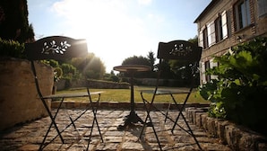 An outdoor seating area with stone flooring and metal chairs, surrounded by greenery and a building in the background.