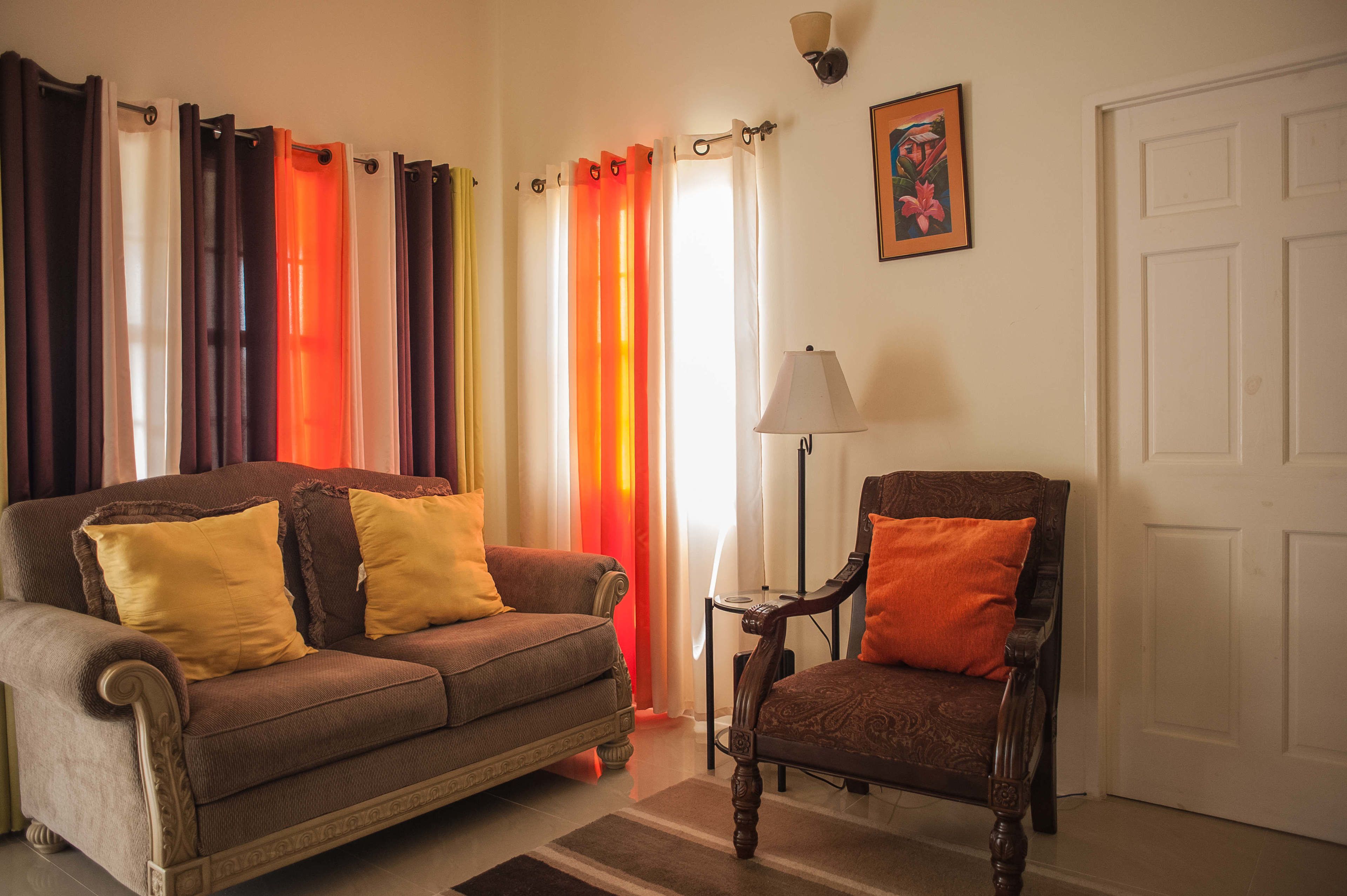 A living room with a brown sofa and a wooden chair, both with cushions. A floor lamp and a painting are on the wall.