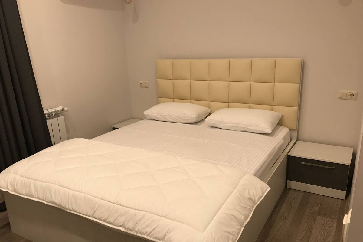 A neatly made bed with a quilted white duvet, a padded headboard, and a bedside table with a drawer.