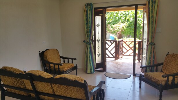 Una habitación con dos sillas de madera con cojines amarillos, una pequeña mesa redonda y un cerramiento de vidrio corredizo que da a un espacio exterior con valla de madera y vegetación.
