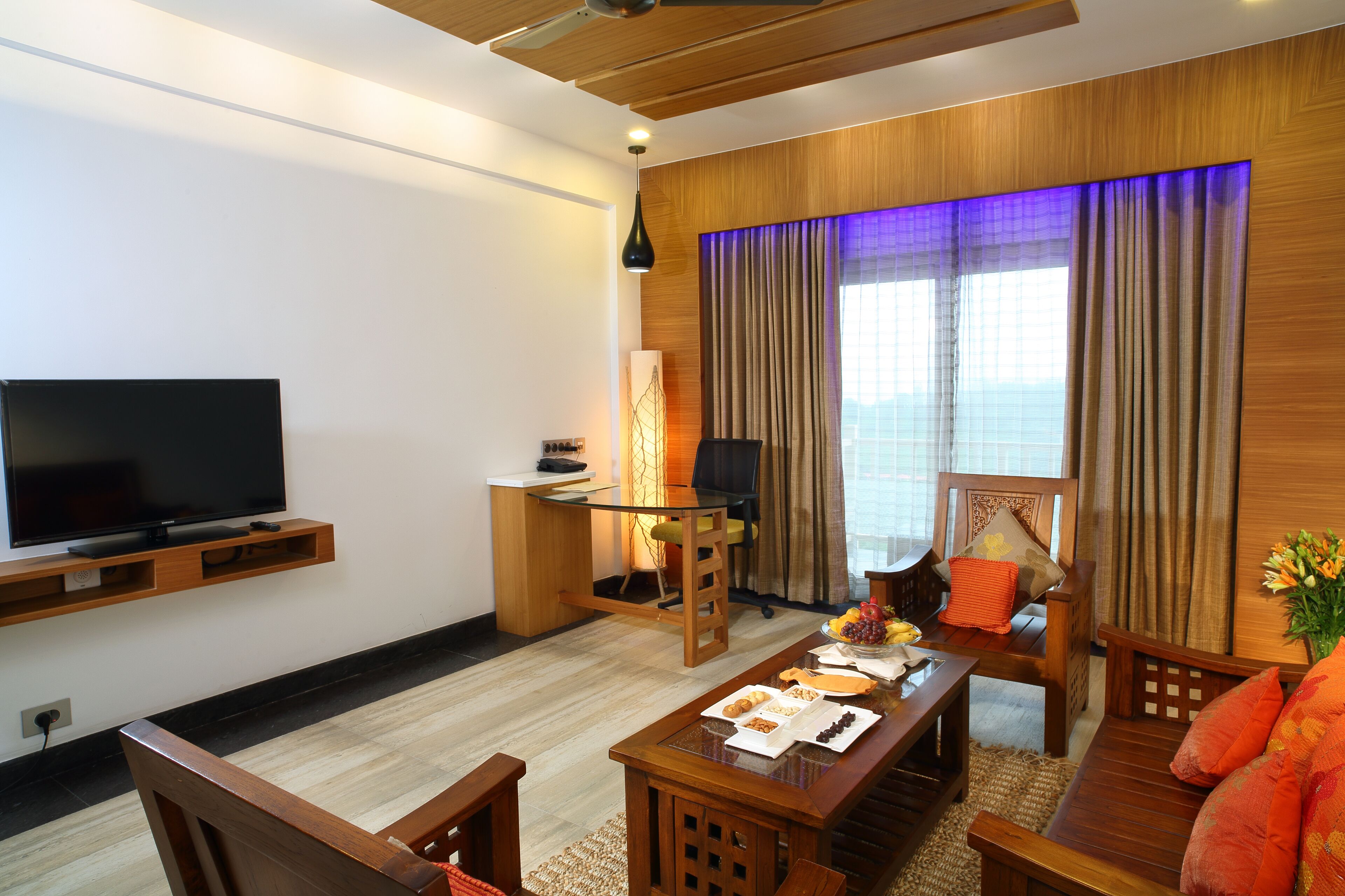 A modern living room with a flat-screen TV, wooden furniture, a dining table with food, and a large window with curtains.