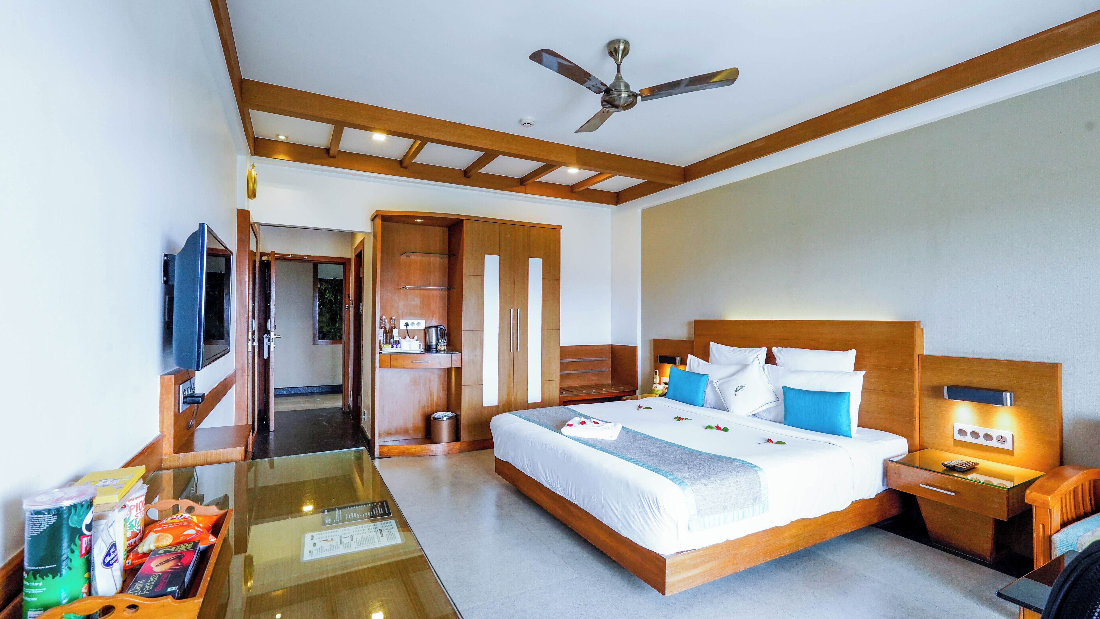 Una habitación de hotel con cama, mesita de noche, ventilador de techo y una mesa de vidrio con diversos objetos.