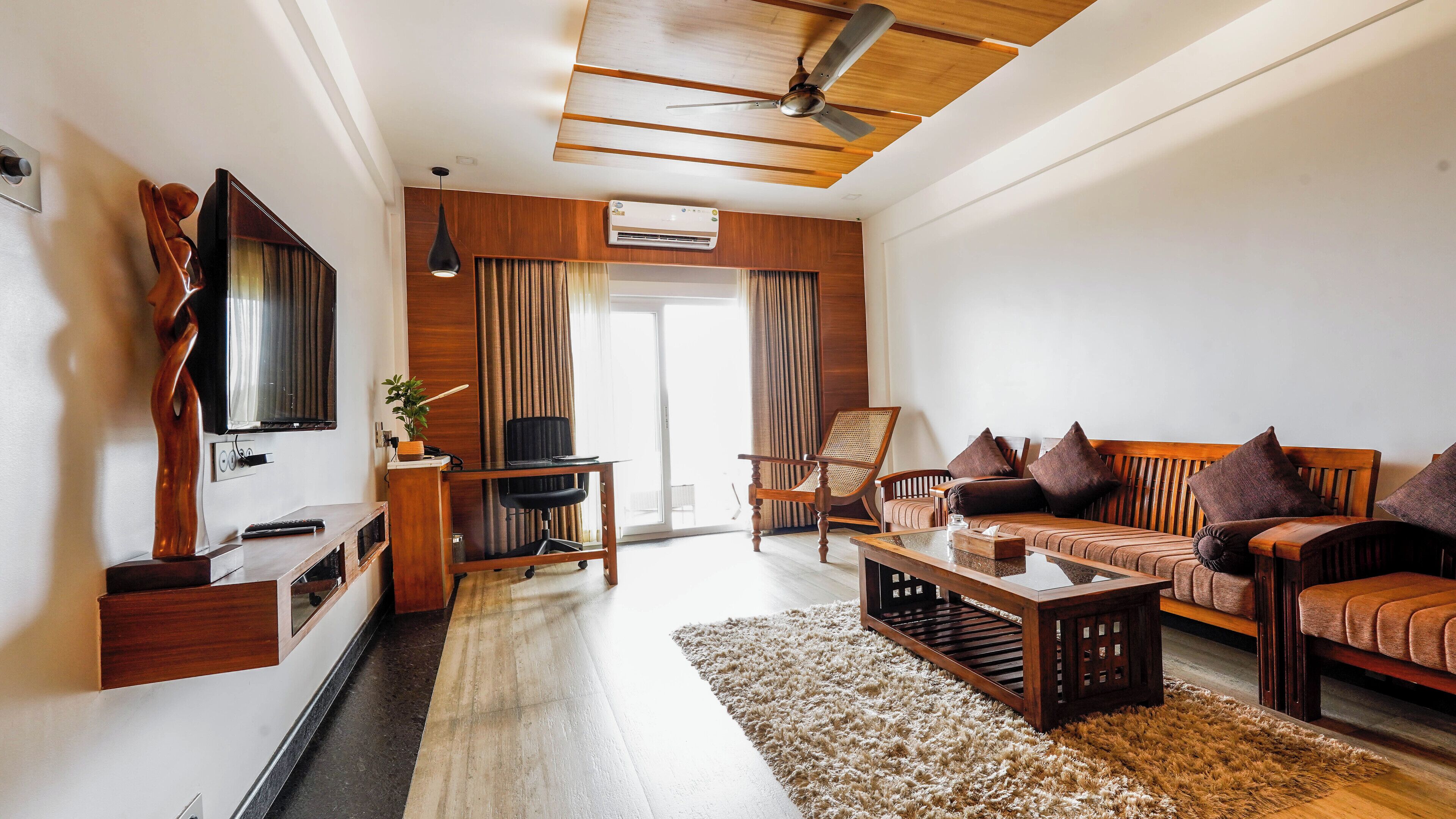 A modern living room with a sofa, armchair, coffee table, and TV. There is a wooden ceiling fan, a ceiling-mounted air conditioner, and a sliding glass door leading to a balcony.