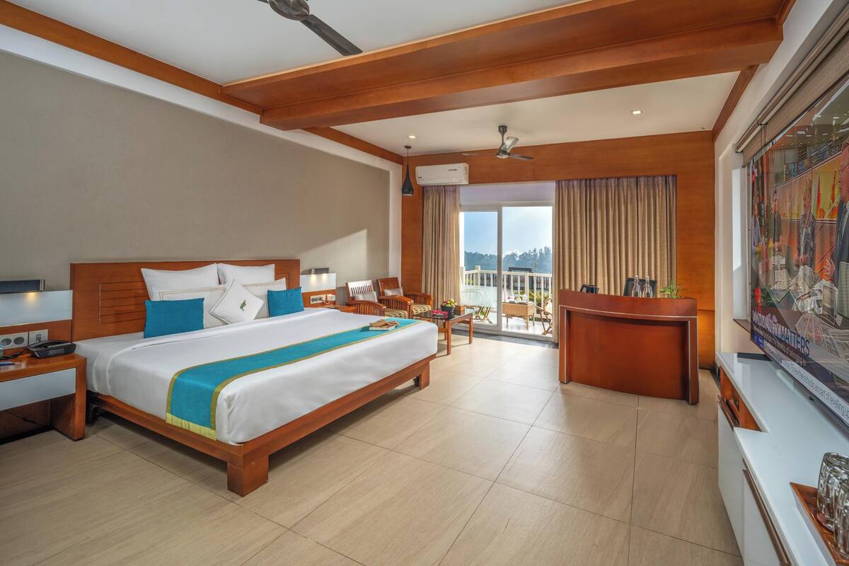 A hotel room with a large bed, a desk, a chair, a balcony with a view, and a ceiling fan.