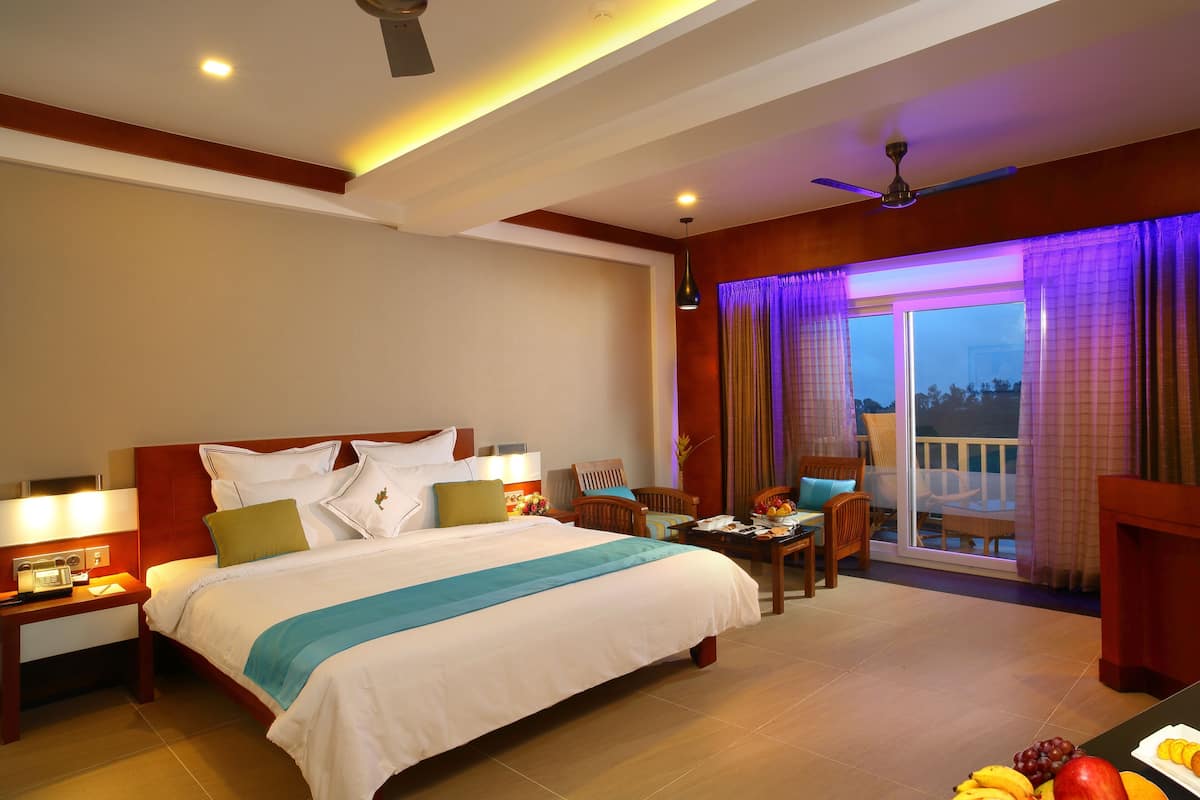 A hotel room with a bed, bedside table, lamp, chair, dining table, fruit, and a balcony with curtains.