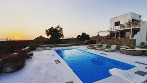 A swimming pool with lounge chairs, a stone wall, and a modern building with a balcony. - Villa Mozaik (Naxos)