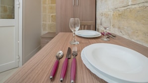 Un tavolo da pranzo apparecchiato con piatti bianchi, posate con manici rosa e bicchieri da vino, con un armadietto in legno e una parete in pietra sullo sfondo.