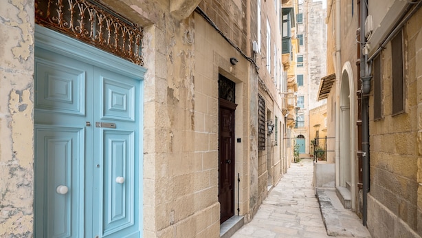 Un stretto vicolo con una porta azzurra a sinistra e una porta marrone scuro a destra. Il vicolo è delimitato da edifici con muri in pietra e un mix di stili architettonici.