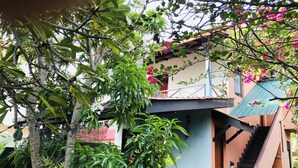 A residential building with a staircase leading to a balcony, surrounded by lush greenery and flowering plants.