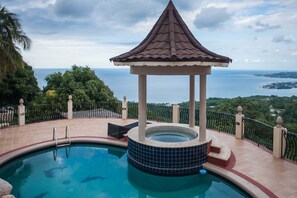 A swimming pool with a gazebo, surrounded by a fence, overlooking a scenic view of the sea and greenery.