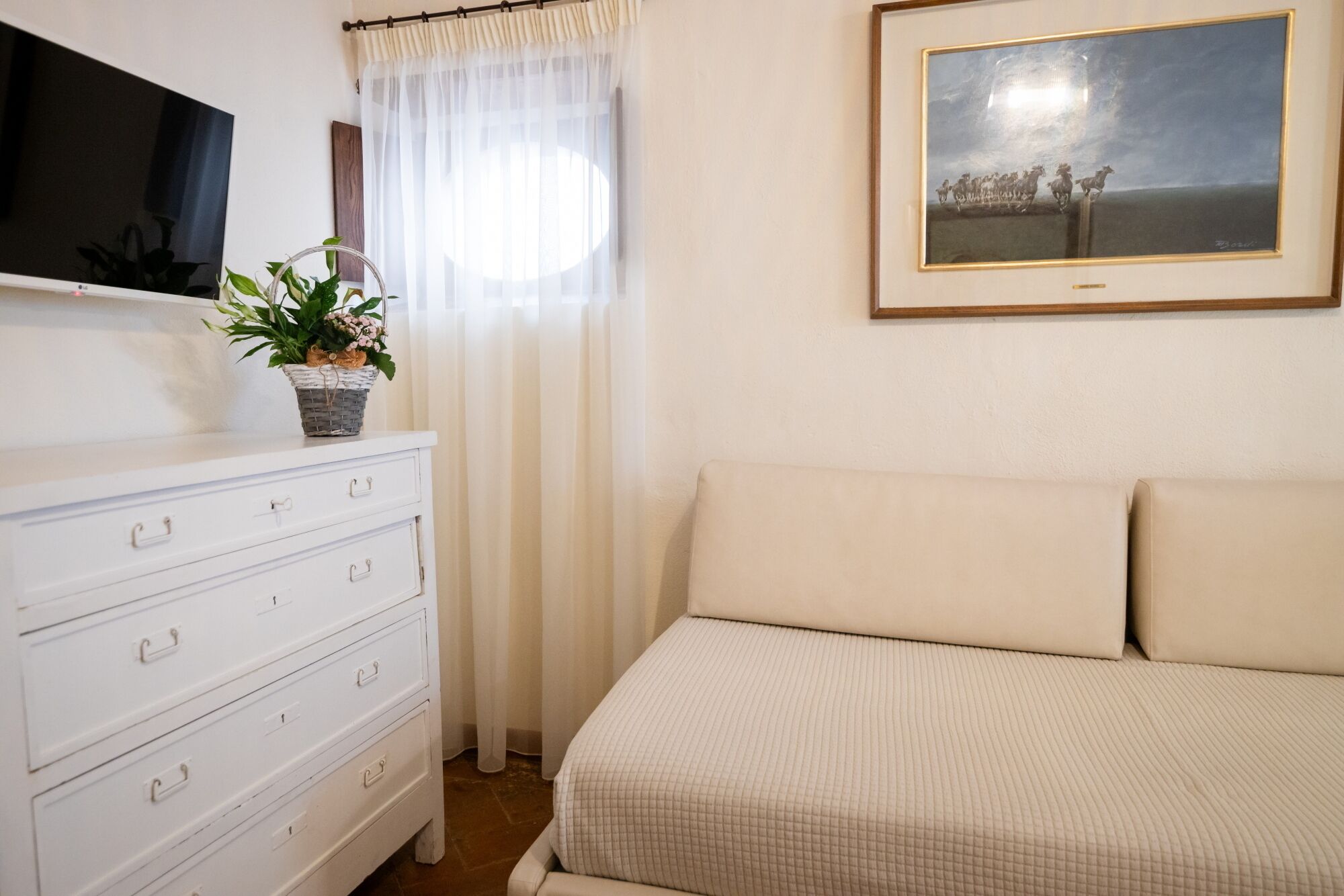 Une chambre à coucher avec un lit, une commode, un téléviseur, une fenêtre avec des rideaux et un tableau au mur.