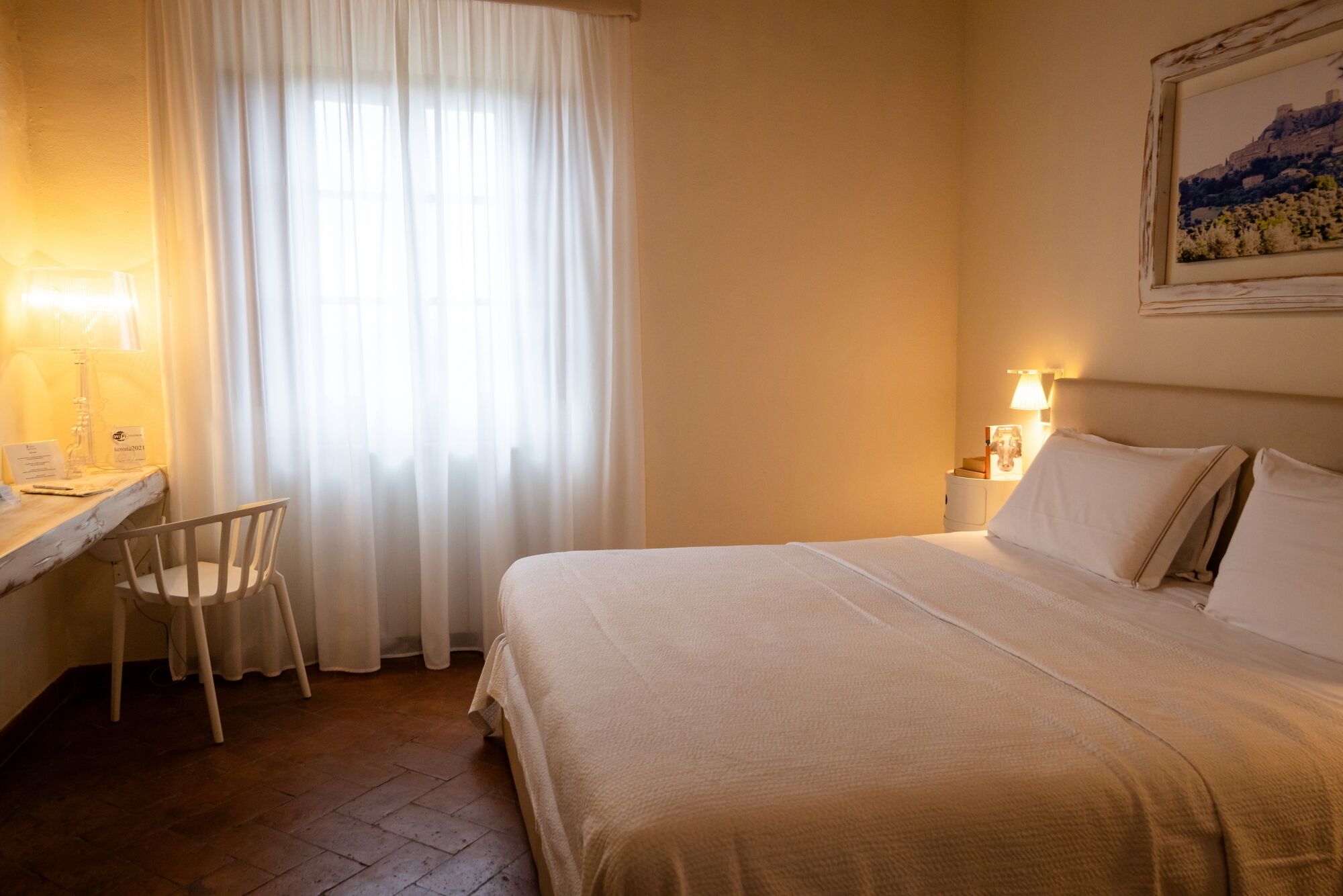 Une chambre d’hôtel comprenant un lit, un bureau avec une chaise, une fenêtre avec des rideaux et une lampe fixée au mur.