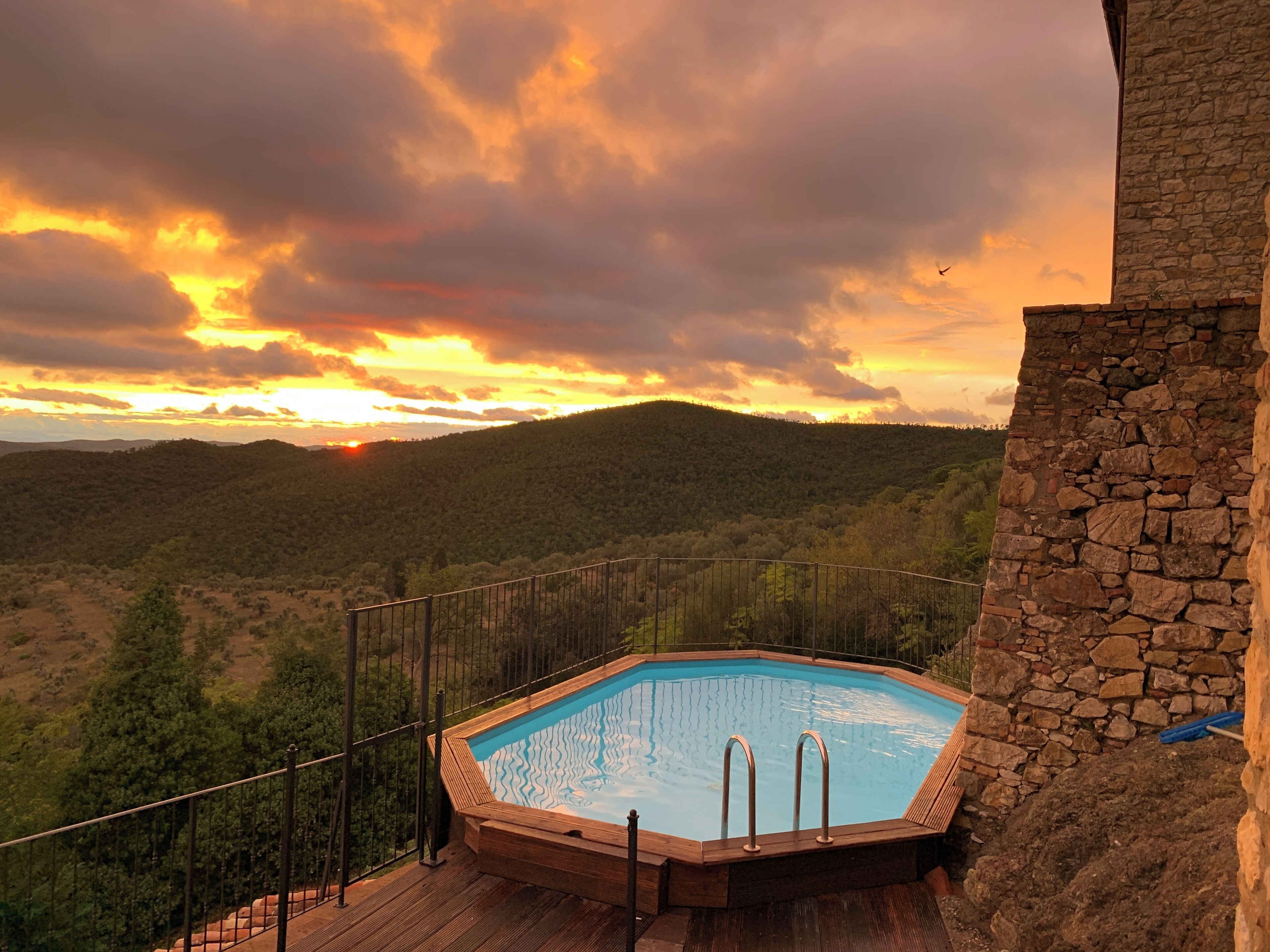 Ein Whirlpool auf einer Holzterrasse mit einer Steinsäule, mit Blick auf eine bewaldete Landschaft bei Sonnenuntergang.