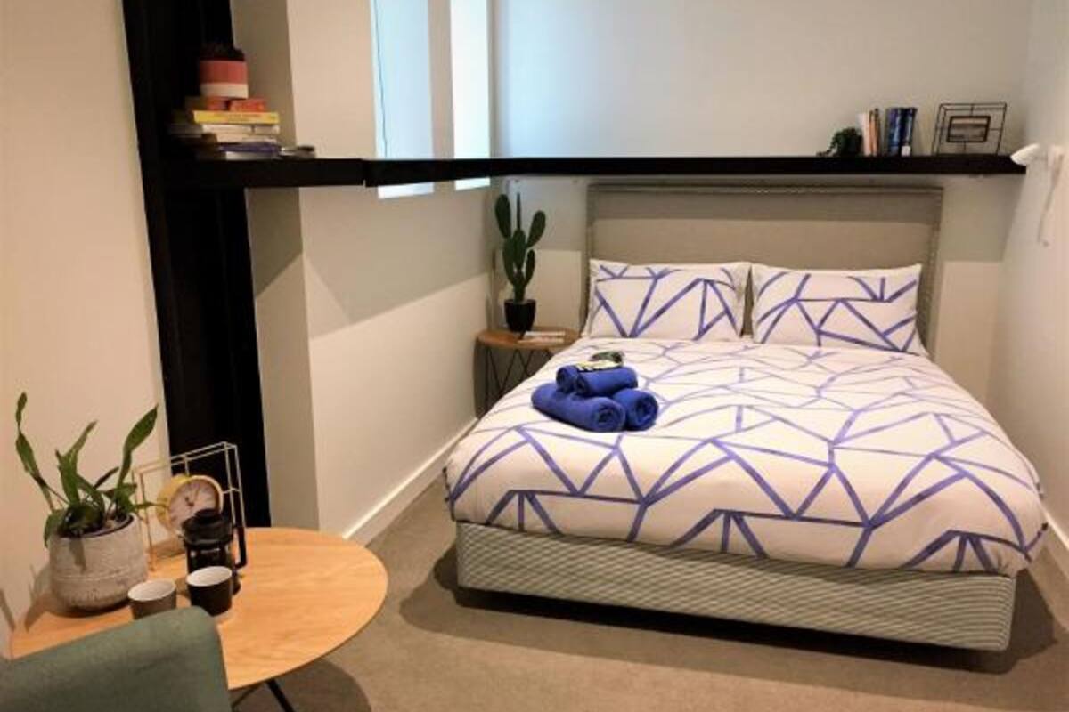 A neatly arranged bedroom with a bed, a small round table, a chair, a shelf with books, and a potted plant.