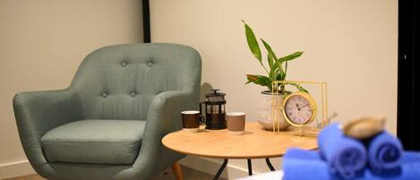 Une pièce avec un fauteuil bleu clair, une petite table ronde sur laquelle se trouvent une cafetière, des tasses, une horloge et un pot de fleurs.