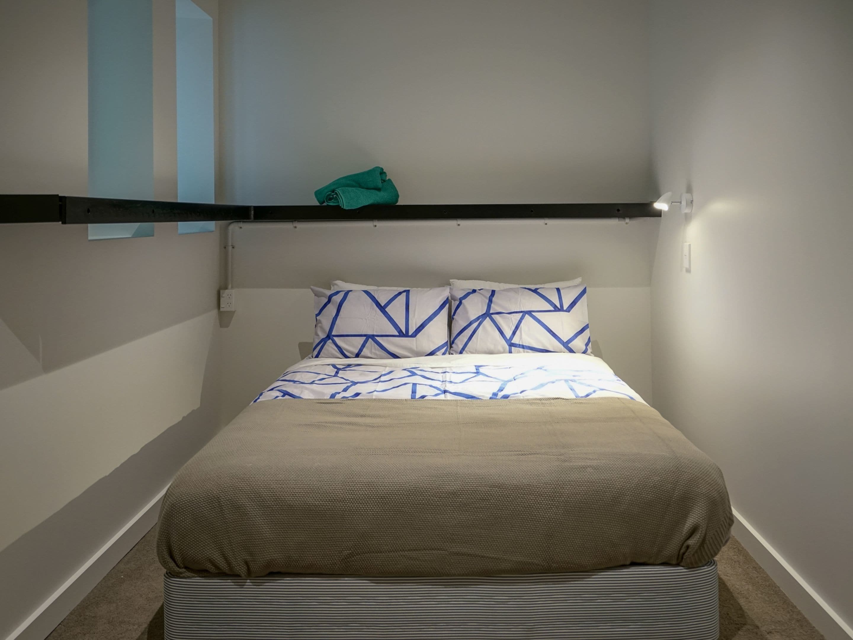A small, neatly arranged bedroom with a single bed, a shelf above the bed, and a wall-mounted light.