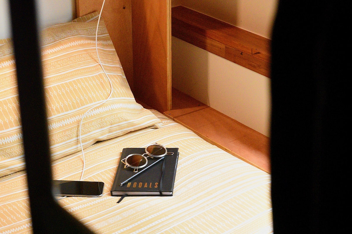 A bed with a patterned bedspread, a smartphone, a notebook, and a pair of sunglasses on it.