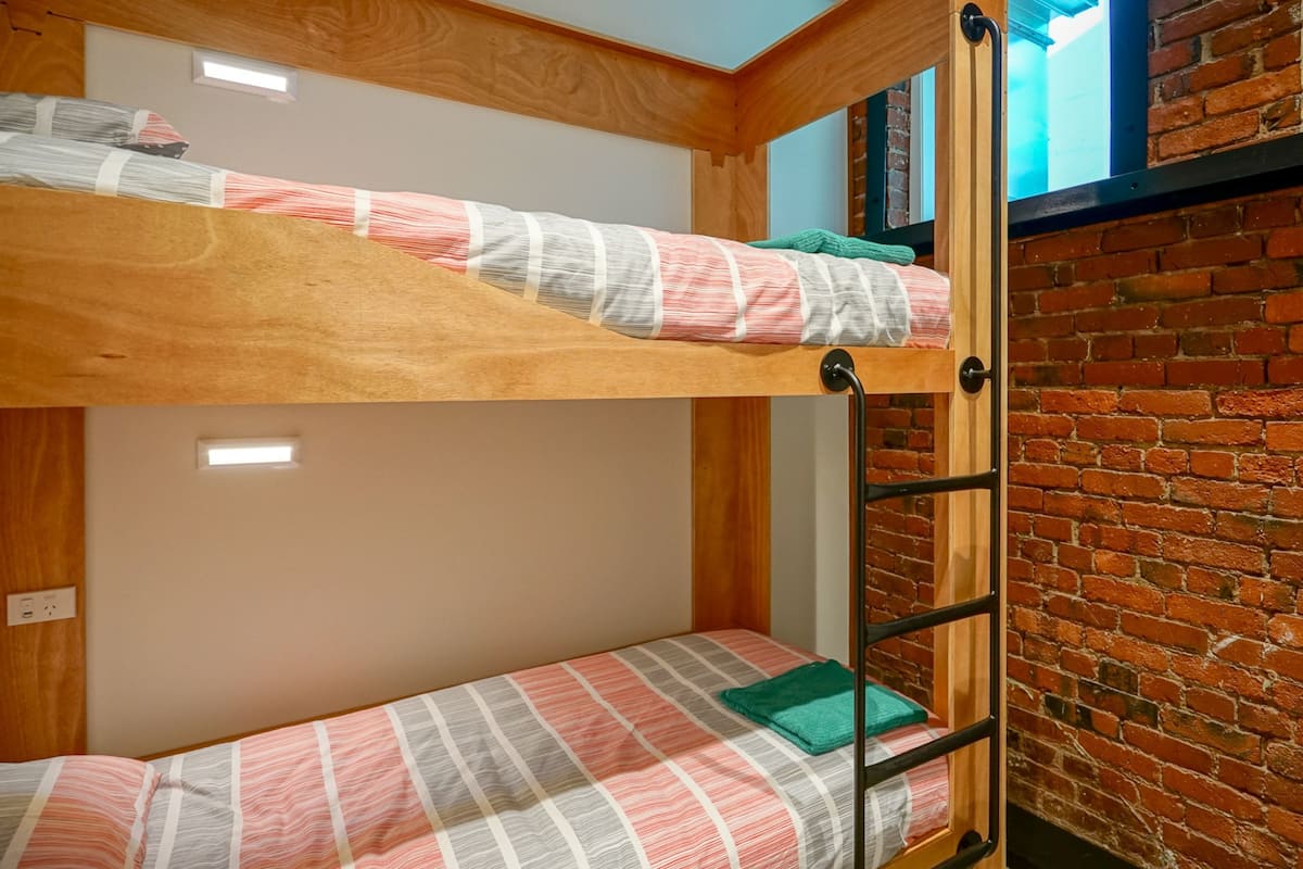 A bunk bed with a wooden headboard, a lower bunk with a plaid blanket, and a brick wall.