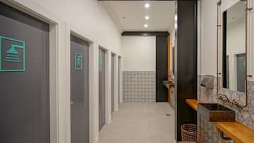 Un pasillo con varias puertas, un muro con baldosas, un lavabo con espejo y un cesto de basura.