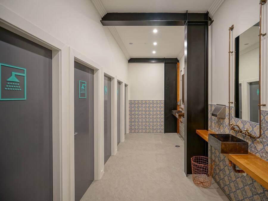 Un pasillo con varias puertas, un muro con baldosas, un lavabo con espejo y un cesto de basura.