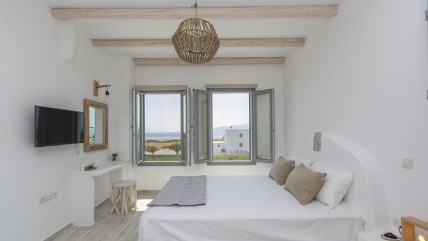 A bedroom with a bed, a TV, a small stool, and a window with an open shutter. - Salinus Villas (Naxos)