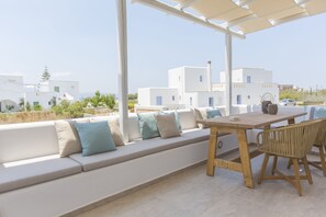 A covered outdoor seating area with a wooden table and chairs, a white sofa with cushions, and a view of white buildings in the background. - Salinus Villas (Naxos)