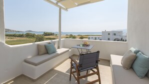 A balcony with a seating area, a table, and a view of the sea and distant buildings. - Salinus Villas (Naxos)