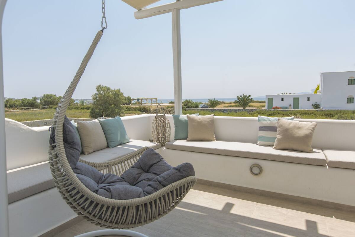 A white outdoor seating area with a hanging chair, cushions, and a view of the sea and buildings.