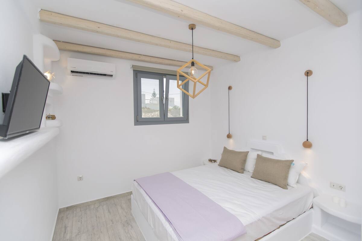 A bedroom with a bed, a window, a TV on a stand, and a hanging light fixture.