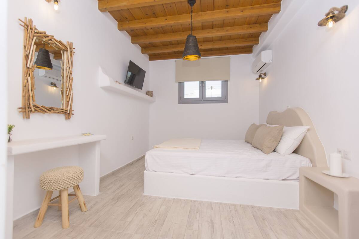 A bedroom with a bed, a wooden ceiling, a TV on a shelf, a window, and a hanging light.
