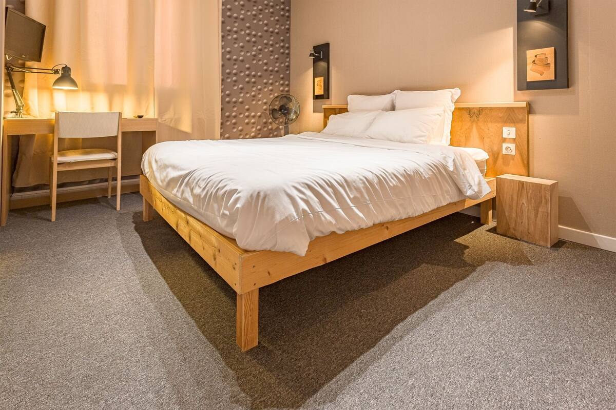 A neatly arranged bedroom with a wooden bed, a desk with a chair, a standing lamp, and a wall-mounted light fixture.