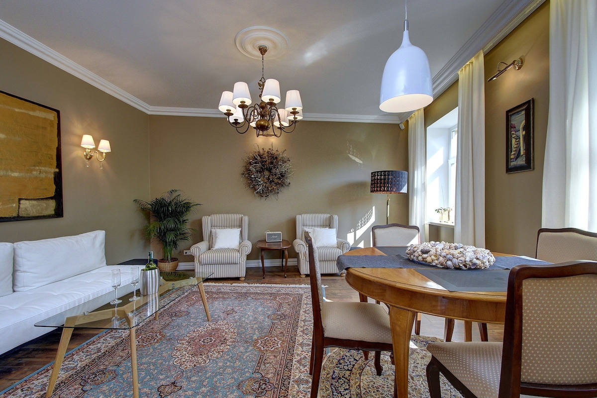 A well-lit living room with a chandelier, a dining table with chairs, a sofa, and a patterned rug.