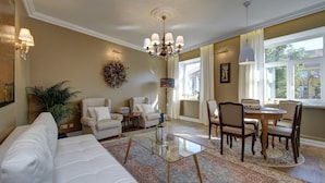 A living room with a white sofa, a glass coffee table, a chandelier, and a dining area with chairs and a table.