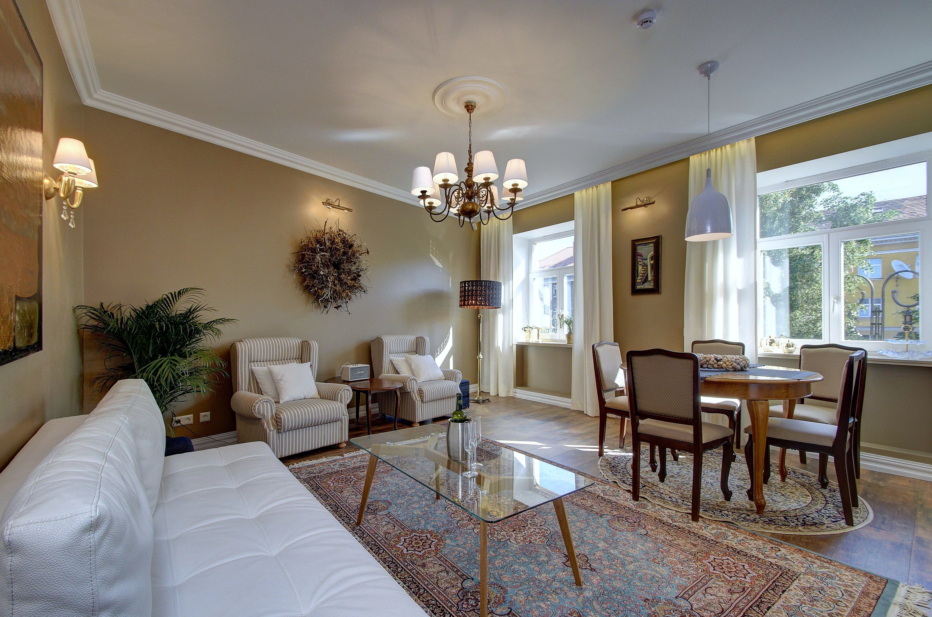 A living room with a sofa, chairs, a glass coffee table, a chandelier, and a view of the outdoors through a window.