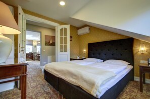 A hotel room with a large bed, a desk with a lamp, a chair, a dining area visible through an open door, and an air conditioning unit on the wall.