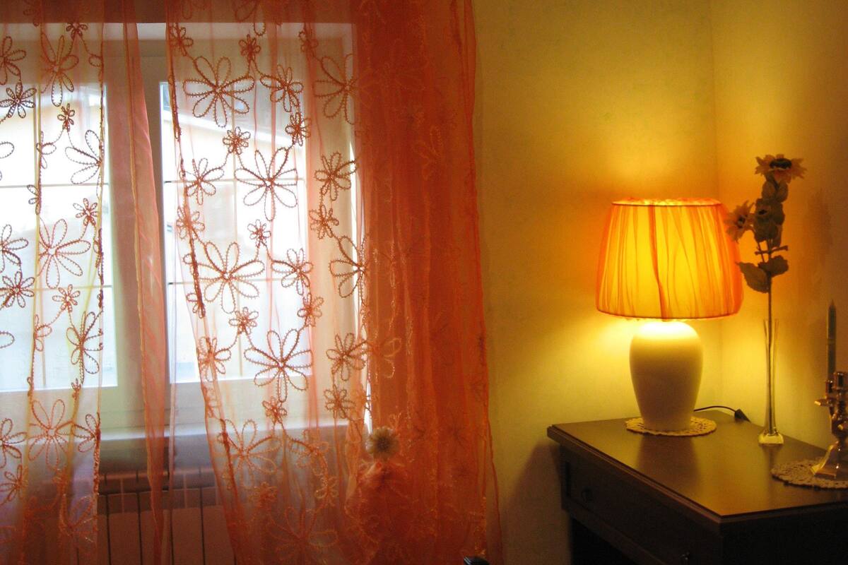 A bedroom with a bed, an air conditioner, a window with a floral curtain, a bedside lamp, and a vase with flowers.