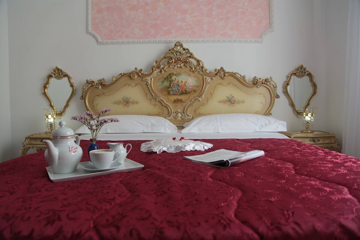 A neatly made bed with a red bedspread, a white tray with a teapot and cups, and a small vase with flowers.