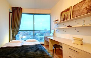 A hotel room with a bed, desk, chair, and a city view through large windows. - Helios City Vilnius Apartments (Vilnius)