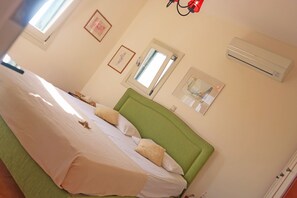 A bedroom with a green headboard, a bed with white linens, two windows, and an air conditioning unit on the wall.