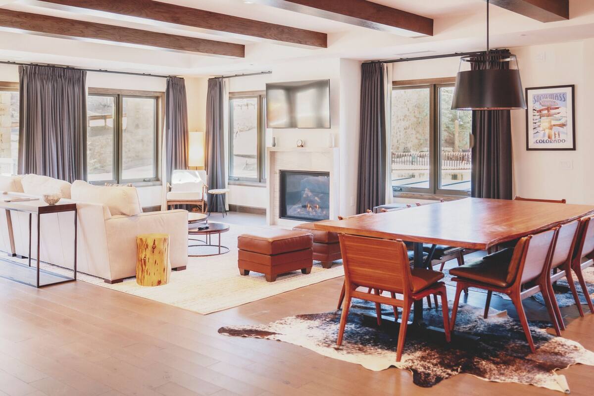Un salon spacieux doté d’une cheminée, un coin repas avec une grande table et des chaises en bois, et un coin salon confortable avec un canapé et des poufs.